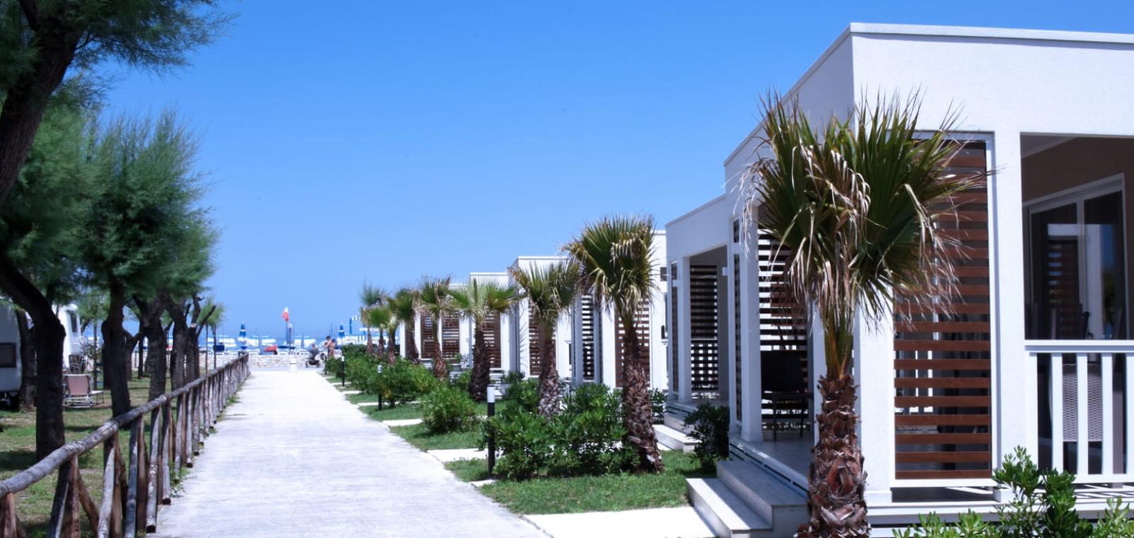 Villini moderni vicino alla spiaggia, circondati da palme e alberi, sotto un cielo azzurro.