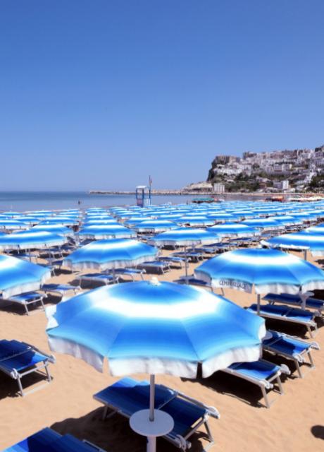 Spiaggia con ombrelloni blu e lettini, mare calmo e città sullo sfondo.