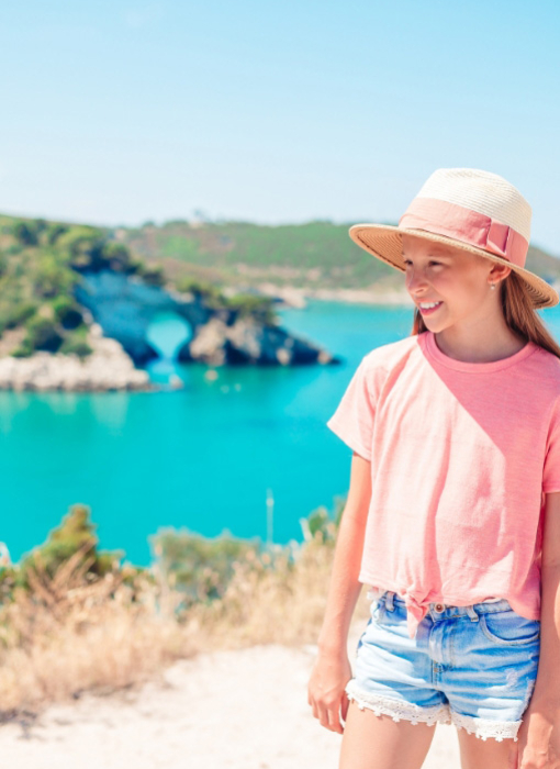 Ragazza sorridente con cappello, in un paesaggio costiero con mare turchese.