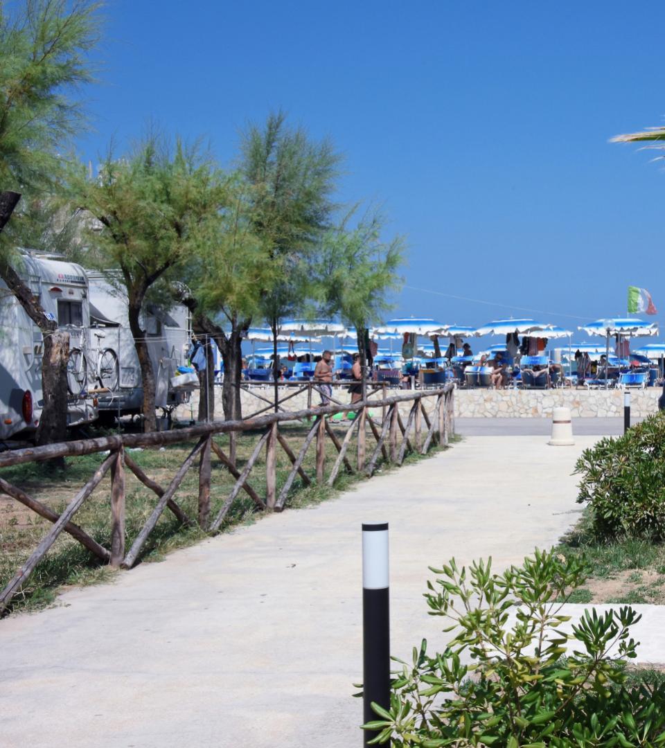 Spiaggia con ombrelloni blu, roulotte e sentiero alberato in una giornata di sole.