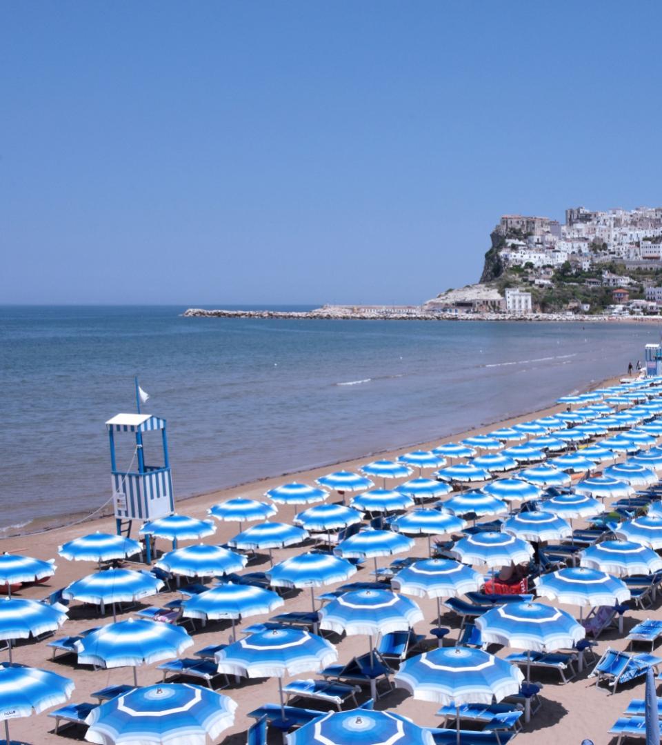 Spiaggia con ombrelloni blu e città sullo sfondo, giornata soleggiata.