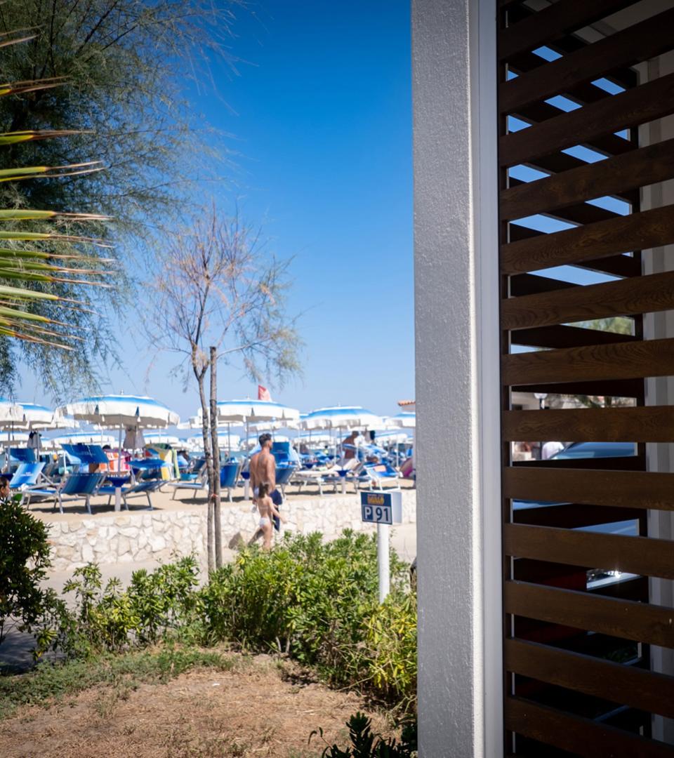 Spiaggia con ombrelloni bianchi e blu, vista da una veranda con persiane in legno.